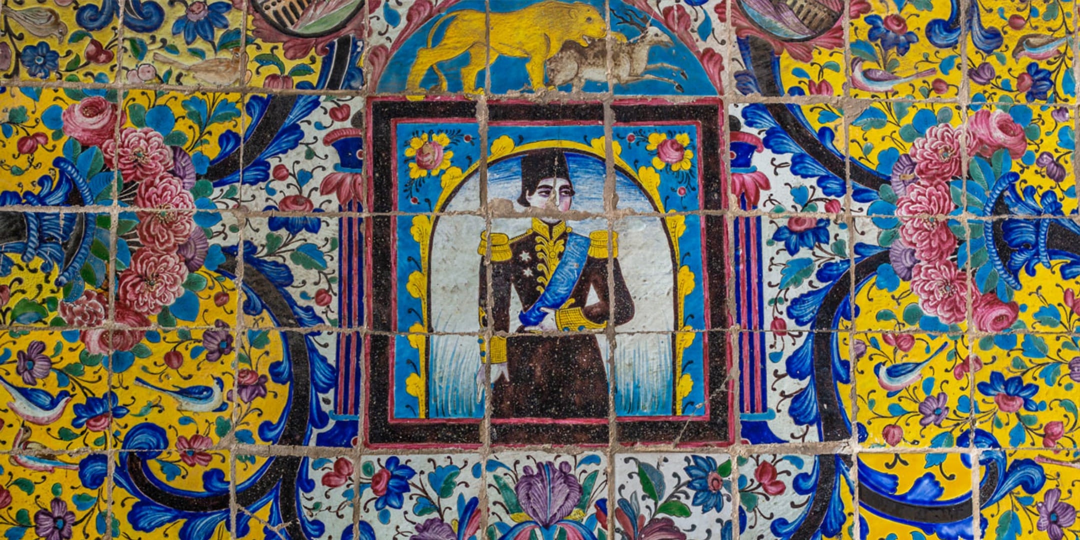 Detailed Persian tilework showing a historic royal figure in uniform, surrounded by floral patterns and a lion motif above, featuring vivid blue, yellow, and pink colors.