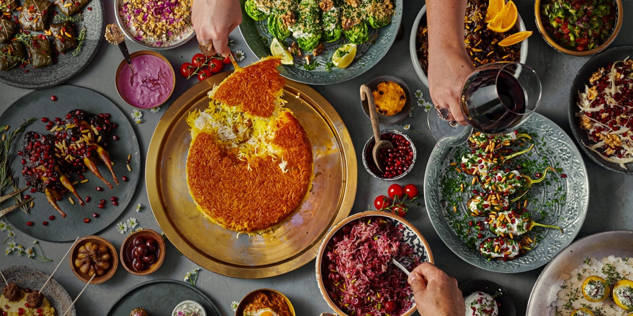 Bird's eye view of a table set with various colorful oriental dishes, rice dish, salads, spices and a glass of red wine held by hands