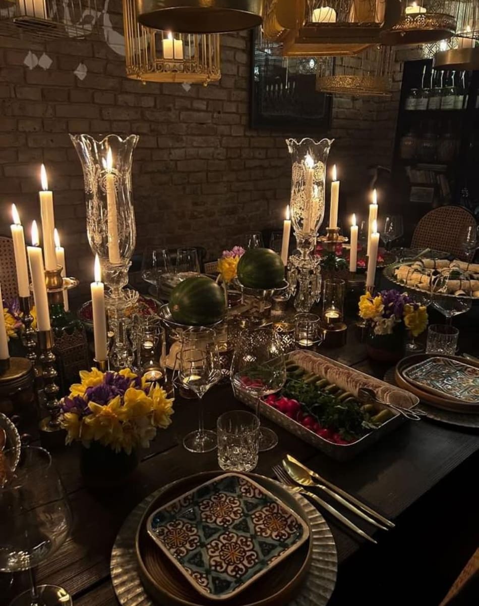 A beautifully set private dining table with lit candles, ornate glassware, fresh yellow and purple flowers, patterned plates, and trays of fresh vegetables, all in a warm, intimate atmosphere with Persian-inspired decor.