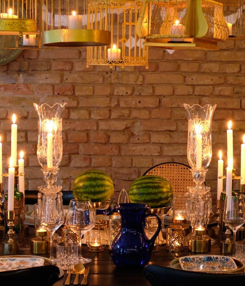 A beautifully set dining table with lit candles, glassware, blue ceramic pitcher, watermelon, and decorative birdcage chandeliers.