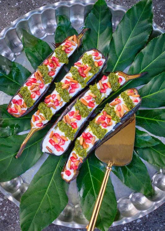 Four grilled eggplant halves with yogurt, pomegranate seeds, pine nuts and green sauce, arranged on large green leaves on a silver tray
