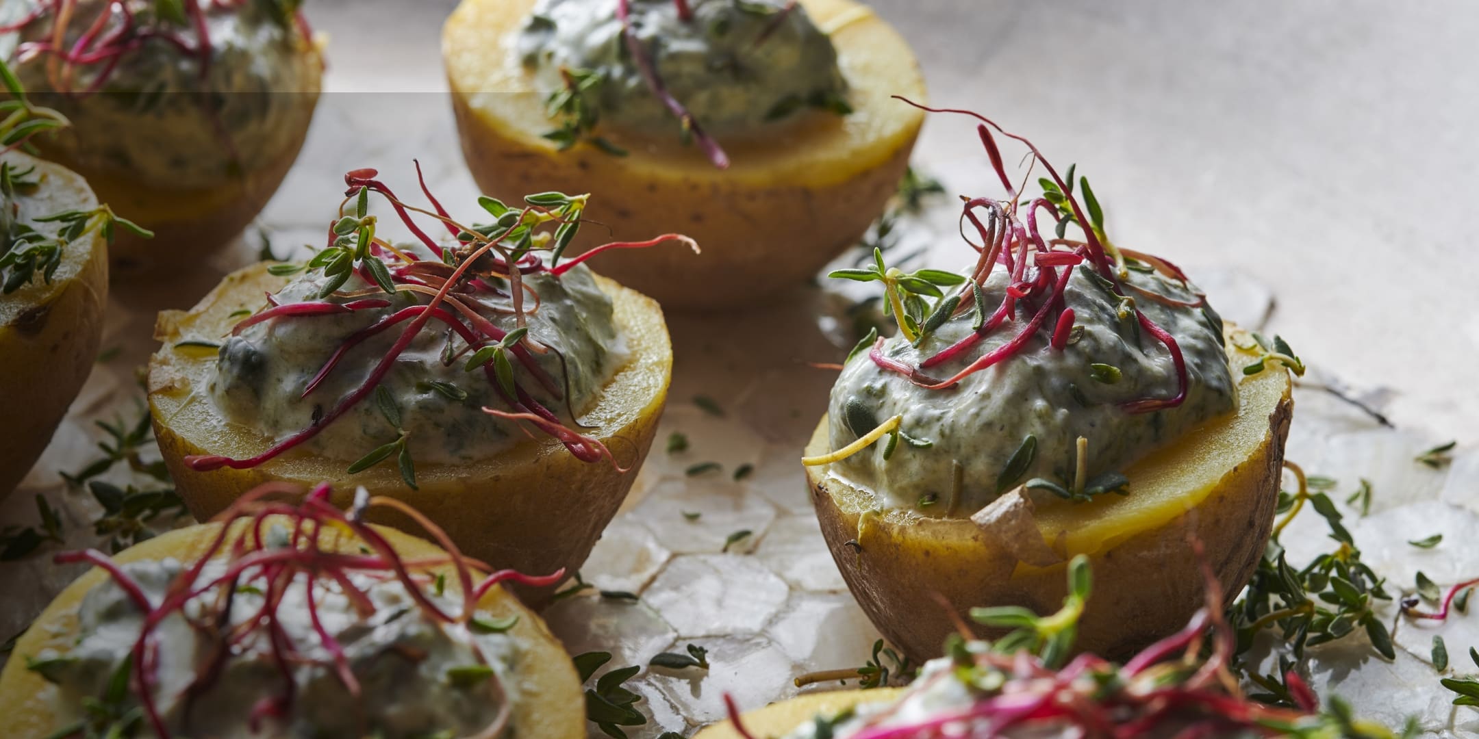 Close-up of halved cooked potatoes topped with creamy yogurt sauce and garnished with fresh herbs and red microgreens on a textured white surface.