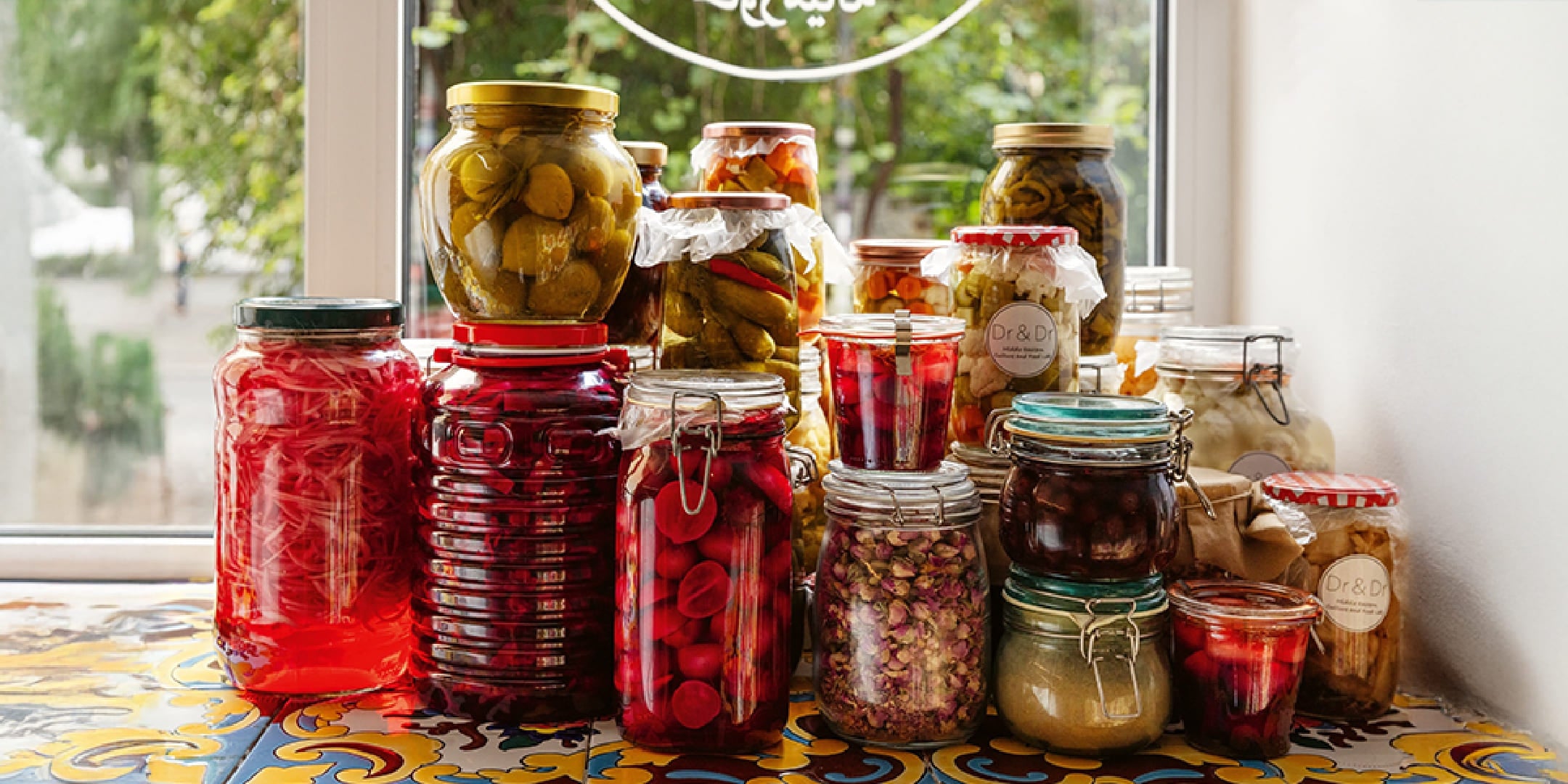 A variety of jars of homemade pickled vegetables, pickles, and herbs on a colorful tile shelf in front of a window, a perfect deli selection for storage.