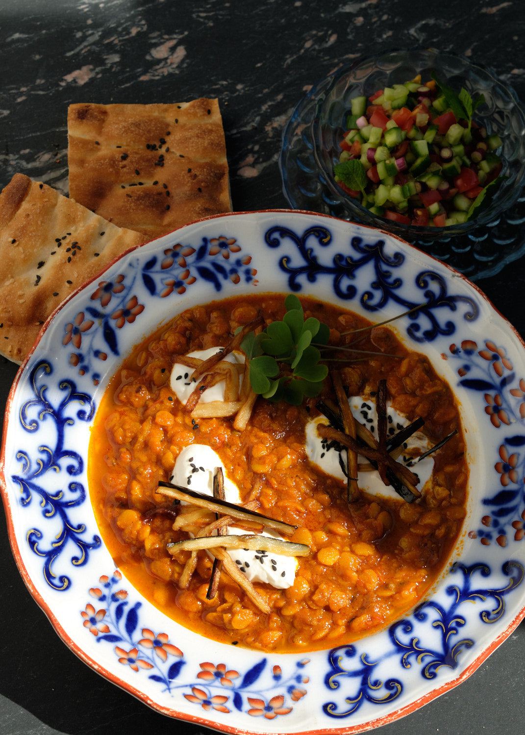 Orientally decorated plate with Persian lentil stew, garnished with yogurt, fried vegetable strips and herbs, next to flatbread and a bowl of cucumber and tomato salad