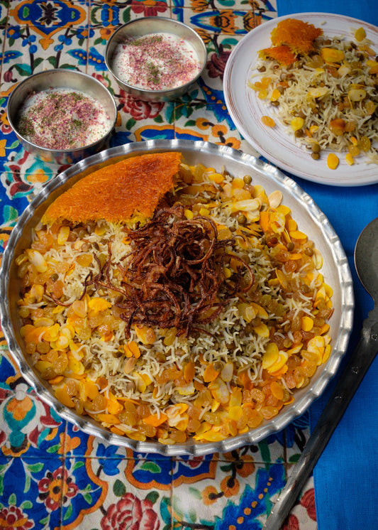 Large bowl with Persian rice dish made of lentils, golden rice crust, raisins, almonds and roasted onions on a colorful oriental cloth, with yogurt dip in metal bowls